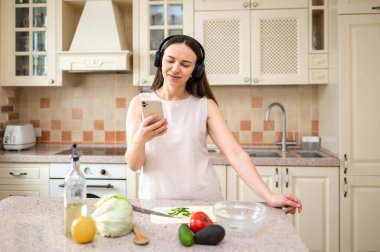  Mutfakta sebze hazırlarken akıllı telefon kullanan bir kadın. Kulaklık takıyor, odaklanmış görünüyor. Tezgahta marul, avokado ve domates gibi taze malzemeler var..