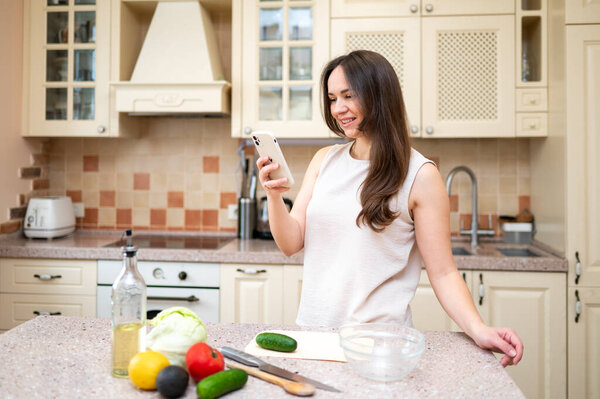 Smiling woman using smartphone while preparing fresh vegetable salad in cozy kitchen. Healthy lifestyle, home cooking and online recipe inspiration