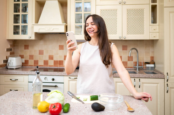 young woman in bright kitchen holding phone and smiling, planning to cook salad. Fresh vegetables and board are on table