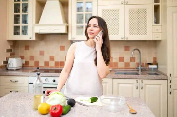 Parlak mutfakta telefonla konuşan, gülümseyen ve salata yapmayı planlayan genç bir kadın. Taze sebzeler ve yemek hazır.