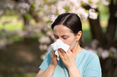 Çiçek açan bahçede dokulara hapşıran kadın, bahar zamanı mevsimsel alerji belirtileri.