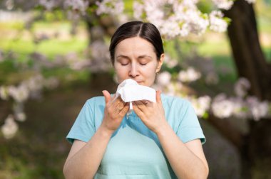 Çiçek açan bahçede dokulara hapşıran kadın, bahar zamanı mevsimsel alerji belirtileri.