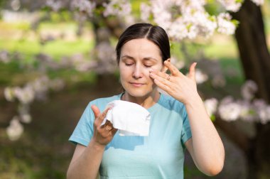 Kadın, çiçek açan bahçede mendile hapşırır. Göz yaşlarını siler. İlkbaharda mevsimsel alerji belirtileri gösterir.