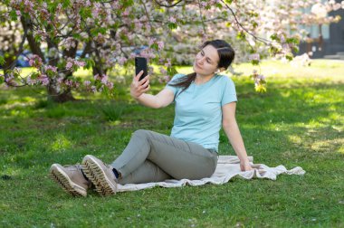 Bahar parkında battaniyeye oturmuş kablosuz kulaklık ve akıllı telefon kullanarak müzik dinleyen genç bir kadın.