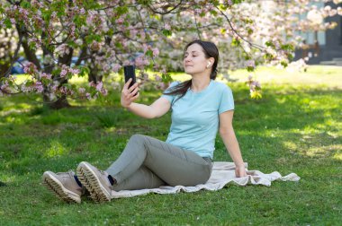 Bahar parkında battaniyeye oturmuş kablosuz kulaklık ve akıllı telefon kullanarak müzik dinleyen genç bir kadın.
