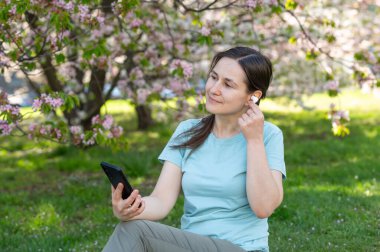 Bahar parkında battaniyeye oturmuş kablosuz kulaklık ve akıllı telefon kullanarak müzik dinleyen genç bir kadın.