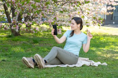 Genç bir kadın lanet bir bahar parkında battaniyeye oturmuş, akıllı telefondan video görüşmesi yapıyor.