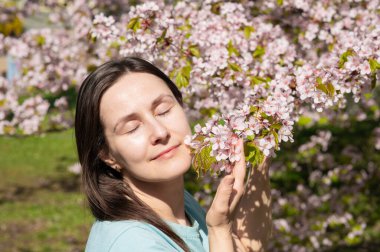 Gözleri kapalı genç bir kadın huzurlu bahar parkında kiraz çiçeklerinin kokusunun tadını çıkarıyor.