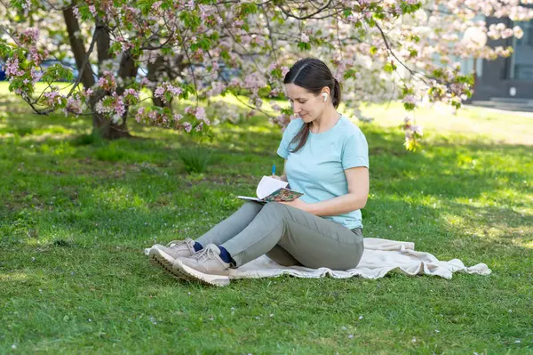 Bir bahar parkında battaniyeye oturmuş not defterine notlar yazan genç bir kadın.