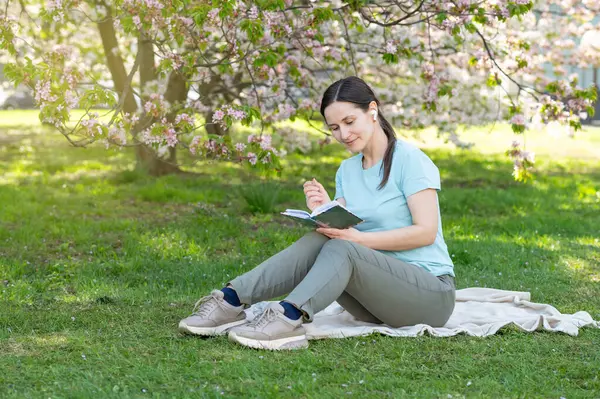 Bir bahar parkında battaniyeye oturmuş not defterine notlar yazan genç bir kadın.