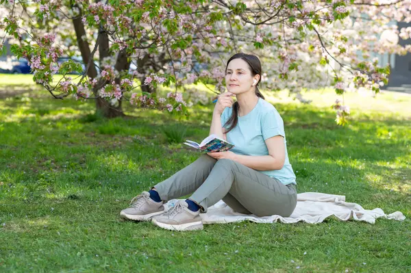 Bir bahar parkında battaniyeye oturmuş not defterine notlar yazan genç bir kadın.