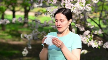 Genç bir kadın hapşırıyor ve çiçek açan sakura bahçesinde mendille gözyaşlarını siliyor. Mevsimsel alerji ve saman nezlesi belirtileri gösteriyor.