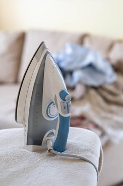 Ütü masasında elektrikli buhar ütüsü, arka planda bulanık kıyafetler ve rahat kanepe, ev işleri konsepti, doğal gün ışığı.