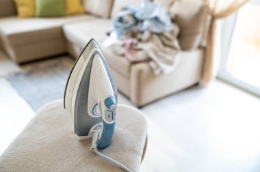 Ütü masasında elektrikli buhar ütüsü, sıcak güneşli bir oturma odasındaki kanepede kırışık çamaşır yığını.