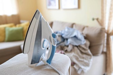 Ütü masasında elektrikli buhar ütüsü ve arka plandaki kanepede kırışık çamaşır yığını.