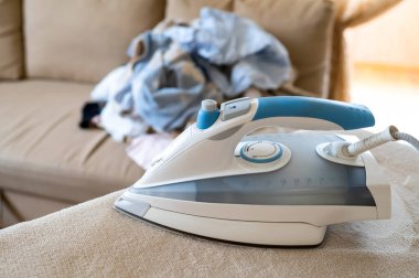 Modern buhar ütüsü kullanıma hazır, arka plan oturma odasında ütülenmemiş çamaşır yığınını gösteriyor.