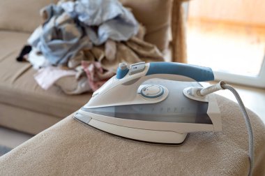 Modern buhar ütüsü kullanıma hazır, arka plan oturma odasında ütülenmemiş çamaşır yığınını gösteriyor.