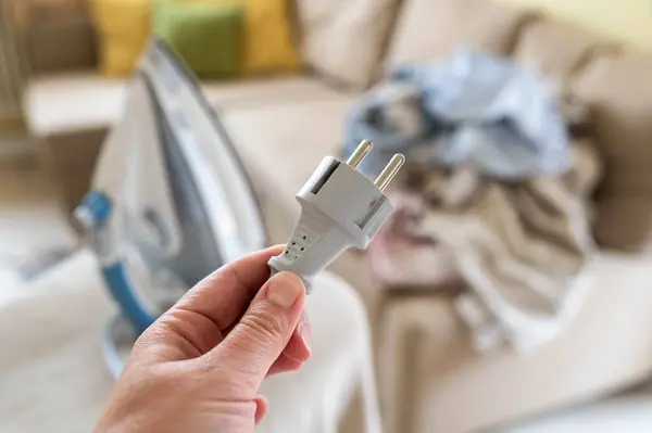 Bir kadının elindeki bağlantısız bir ütüden kopmuş bir elektrik kablosuyla ütü masasının arka planına bağlı bir fiş ve buruşmuş bir çamaşır yığını.