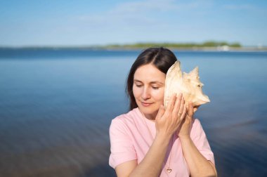 Genç bir kadın göl kenarında deniz kabuğu dinler, gözleri kapalıdır, denizi huzurlu bir şekilde hayal eder.