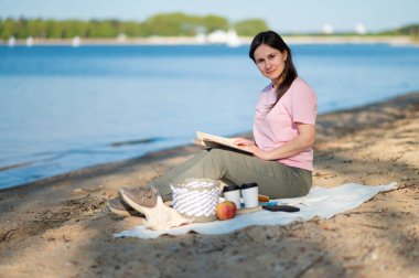 Genç kadın, güneşli göl kıyısında piknik yaparken battaniyede kitap okuyor. Doğanın ve yalnızlığın tadını çıkarıyor.