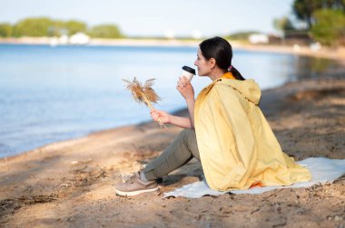 Bir kadın gölün kıyısında oturuyor, kağıt bardaktan sıcak bir içecek içiyor ve elinde kurumuş bir çiçek tutuyordu..