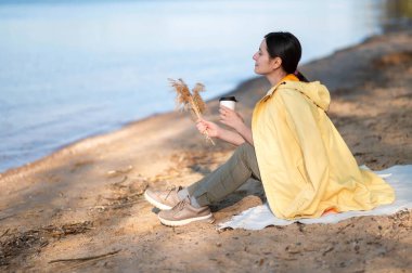 Bir kadın gölün kıyısında oturuyor, kağıt bardaktan sıcak bir içecek içiyor ve elinde kurumuş bir çiçek tutuyordu..