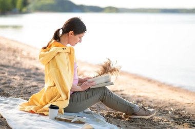 Bir kadın göl kıyısında oturuyor, kitap okuyor ve elinde kurumuş çiçekler tutuyor..