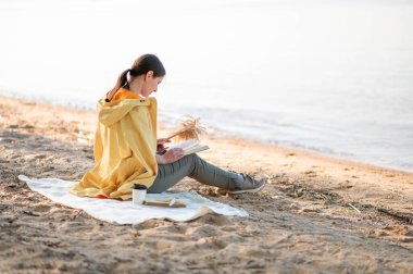 Bir kadın göl kıyısında oturuyor, kitap okuyor ve elinde kurumuş çiçekler tutuyor..