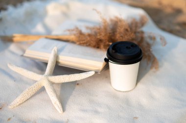 Açık bir kitap, kurumuş çiçekler, sıcak bir içecek ve sarı kumlara yayılmış bir deniz yıldızı..