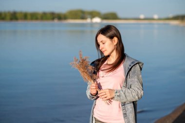 Bir kadın elinde kurumuş çiçeklerle gölün kenarında duruyor, güneşin altında güneşleniyor ve doğada huzurlu bir anın tadını çıkarıyor.