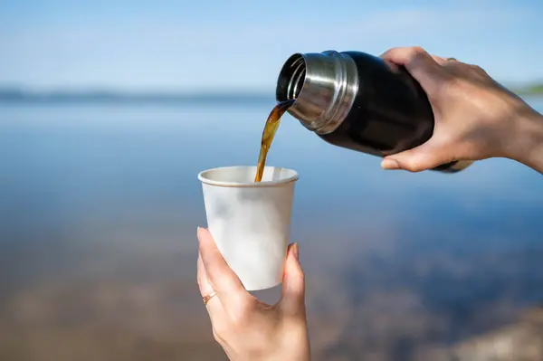 Kadın elleri, güneşli bir günde arka planda manzaralı bir gölle termostan kağıt bardağa sıcak kahve döker.