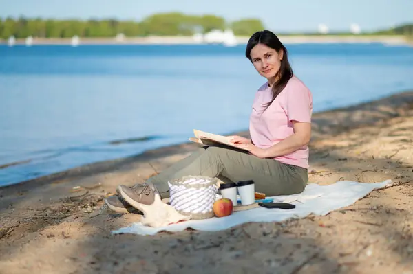 Genç kadın, güneşli göl kıyısında piknik yaparken battaniyede kitap okuyor. Doğanın ve yalnızlığın tadını çıkarıyor.