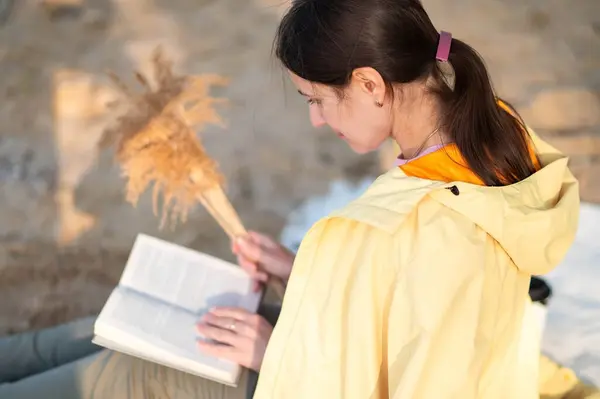 Bir kadın göl kıyısında oturuyor, kitap okuyor ve elinde kurumuş çiçekler tutuyor..