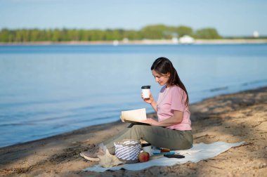 Kitap okuyan ve sıcak bir şeyler içen genç bir kadın güneşli göl kıyısında piknik yaparken battaniyeye oturmuş.