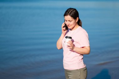 Genç bir kadın güneşli bir günde gölün kıyısında kahve içiyor ve akıllı bir telefonla konuşuyor..