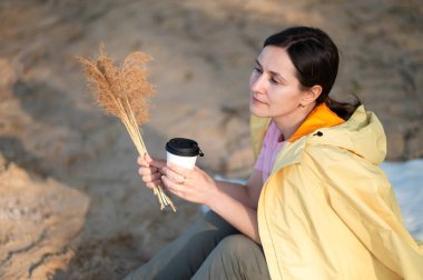 Bir kadın gölün kıyısında oturuyor, kağıt bardaktan sıcak bir içecek içiyor ve elinde kurumuş bir çiçek tutuyordu..