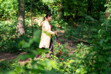 Aktif orta yaşlı kadın yaz ormanlarında İskandinav kutuplarıyla yürüyor, sağlıklı yaşam ve fiziksel refahı teşvik ediyor.