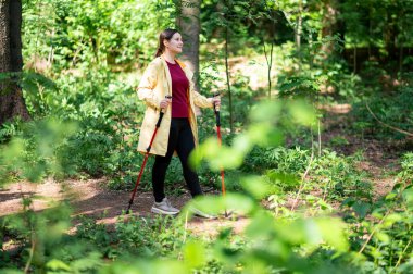 Güneşli bir orman yolunda özel direklerle yürüyen bir kadın aktif yaşam tarzının ve temiz havanın tadını çıkarıyor.