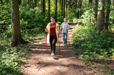 Anne ve oğlu güneşli bir ormanda yürüyor, dışarıda İskandinav yürüyüşü yapıyorlar.