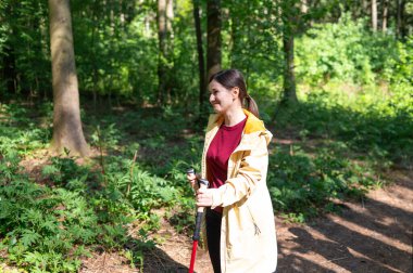 Genç bir kadın güneşli bir ormanda yürüyor, direkle İskandinav yürüyüşü yapıyor.