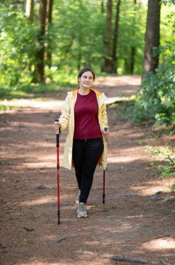 Mutlu kadın gün ışığında ormanda yürüyor, İskandinav yürüyüşü yapıyor ve dışarıda egzersiz yapıyor.