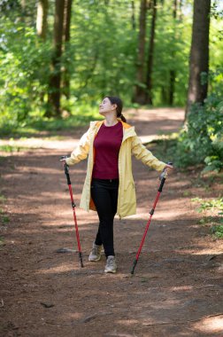 Mutlu kadın gün ışığında ormanda yürüyor, İskandinav yürüyüşü yapıyor ve dışarıda egzersiz yapıyor.