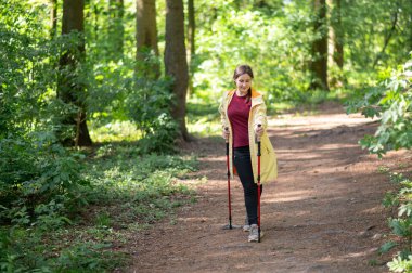 Neşeli bir kadın, güneşli bir orman patikasında sırıklarla yürüme pratiği yapıyor sağlıklı bir yaşam tarzı için zindelik ve doğayı birleştiriyor.