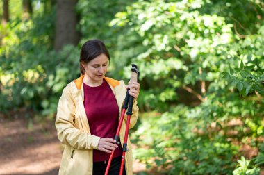 Ormanda yürüyen bir kadın, İskandinav yürüyüş direklerini inceliyor, doğanın tadını çıkarıyor ve aktif bir açık hava egzersizi için hazırlanıyor.