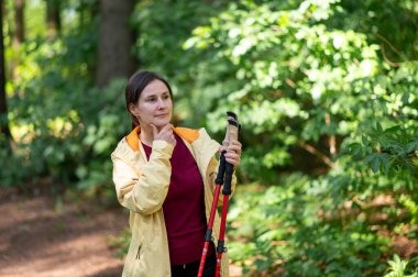 Ormanda yürüyen bir kadın, İskandinav yürüyüş direklerini inceliyor, doğanın tadını çıkarıyor ve aktif bir açık hava egzersizi için hazırlanıyor.