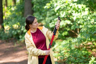 Ormanda yürüyen bir kadın, İskandinav yürüyüş direklerini inceliyor, doğanın tadını çıkarıyor ve aktif bir açık hava egzersizi için hazırlanıyor.