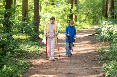 Anne ve 11 yaşındaki oğlu güneşli bir ormanda yürüyüş yapıyor ve İskandinav yürüyüşü yapıyor.