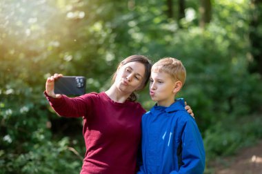 Anne ve oğlu bahar ormanı yürüyüşü sırasında selfie çekerken komik surat ifadeleri takınıyorlar, doğada eğlenceli aile zamanlarının tadını çıkarıyorlar.