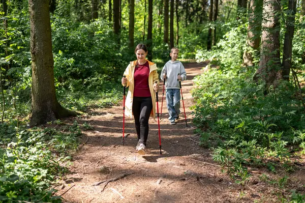 Anne ve oğlu güneşli bir ormanda yürüyor, dışarıda İskandinav yürüyüşü yapıyorlar.