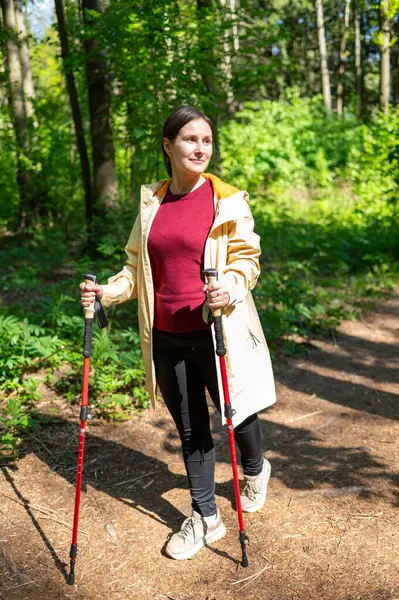 Genç bir kadın güneşli bir ormanda yürüyor, direkle İskandinav yürüyüşü yapıyor.